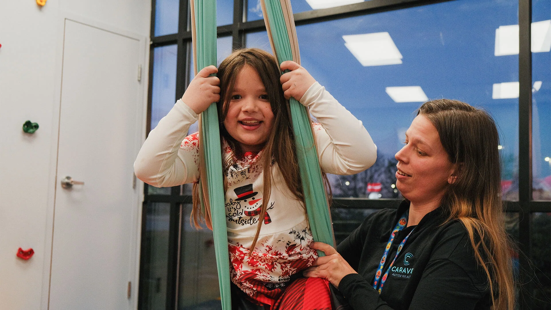 Caregiver assisting a smiling child on an indoor swing in a bright playroom.
