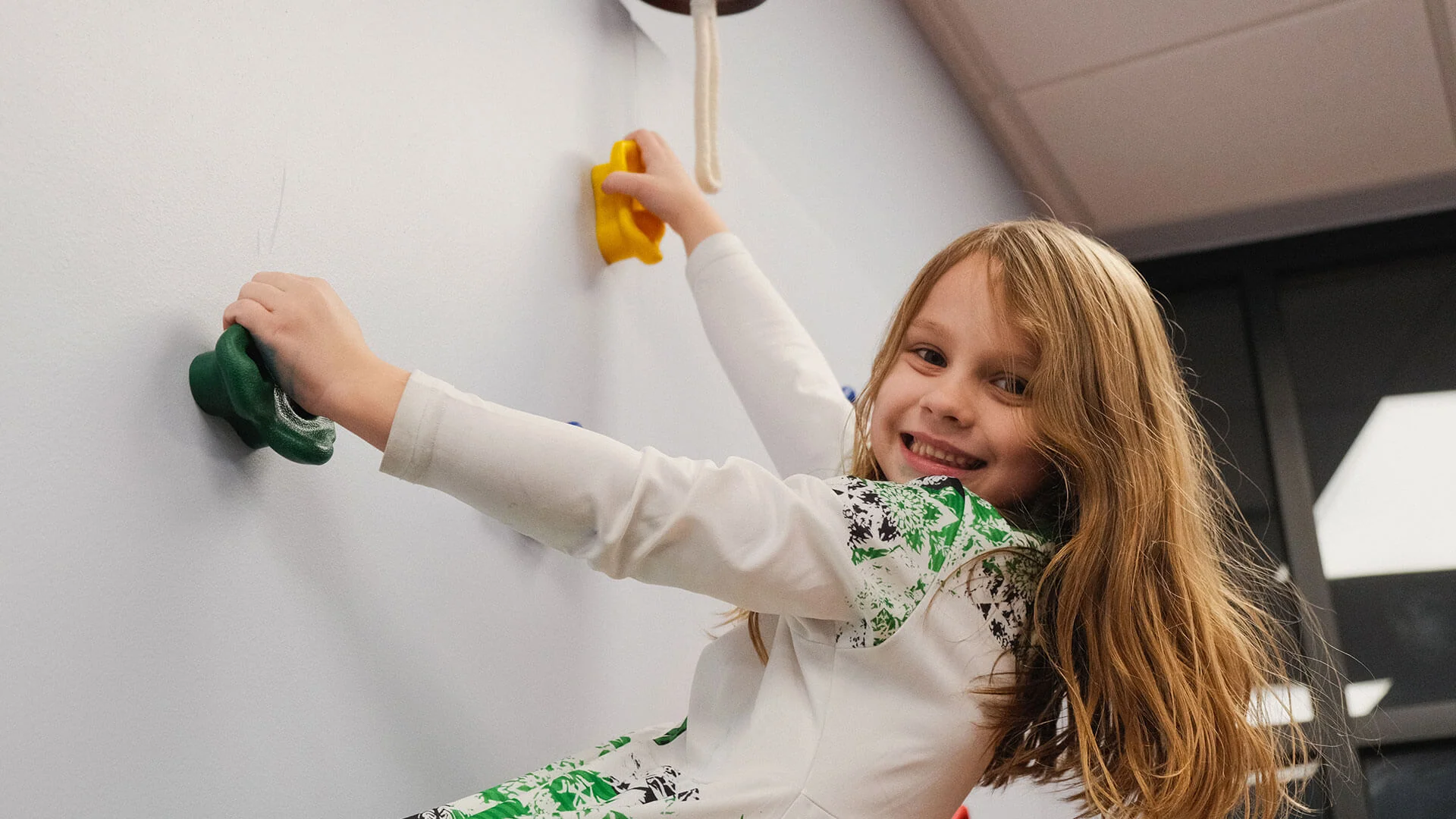 Smiling child climbing an indoor play wall with colorful holds.