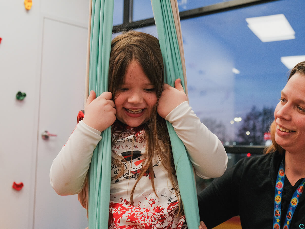 Child in a colorful room enjoying a swing while smiling with a caregiver.