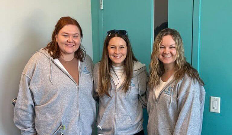 Three women in matching gray hoodies standing in front of a teal door.