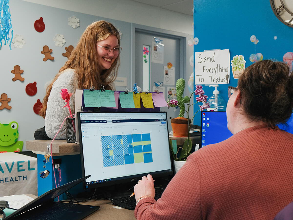 Two women in a community office discussing a scheduling system on a computer screen.