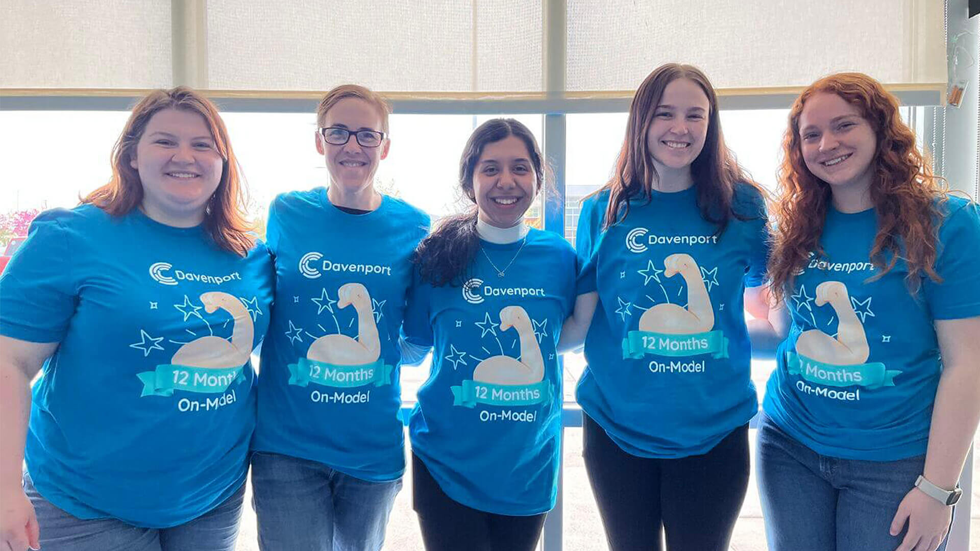 Five people in blue Davenport shirts stand together, smiling in front of large windows.