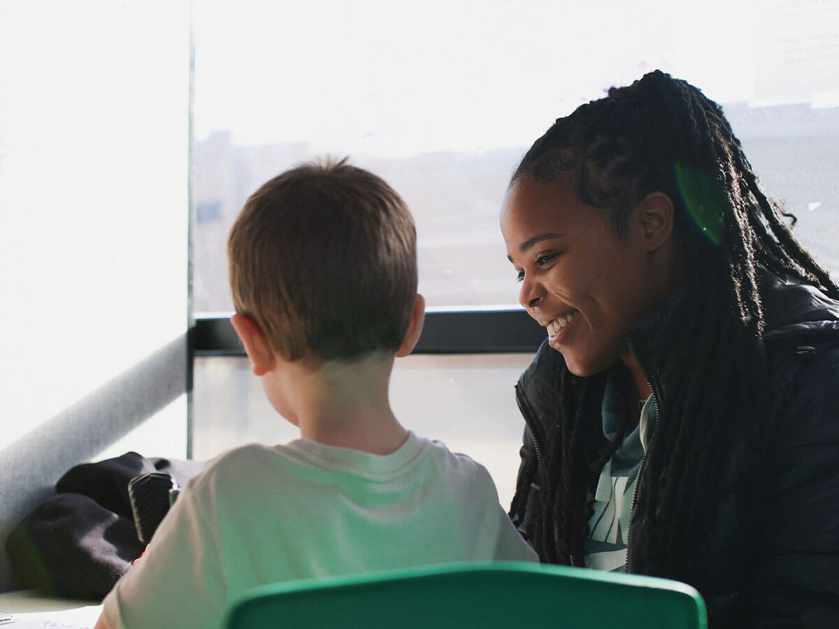 Smiling instructor helps young student in classroom setting.