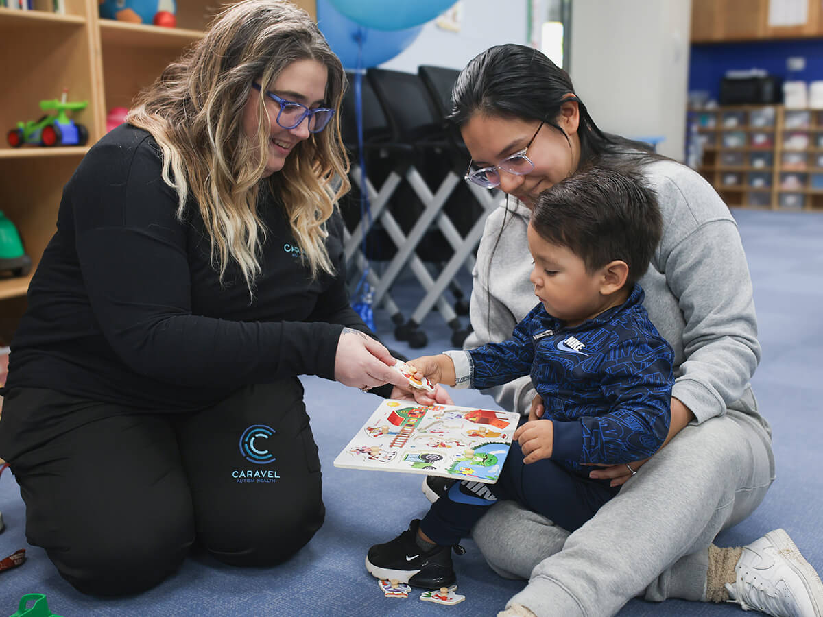 Woman engaging with a child and caregiver during an educational activity in a playroom.