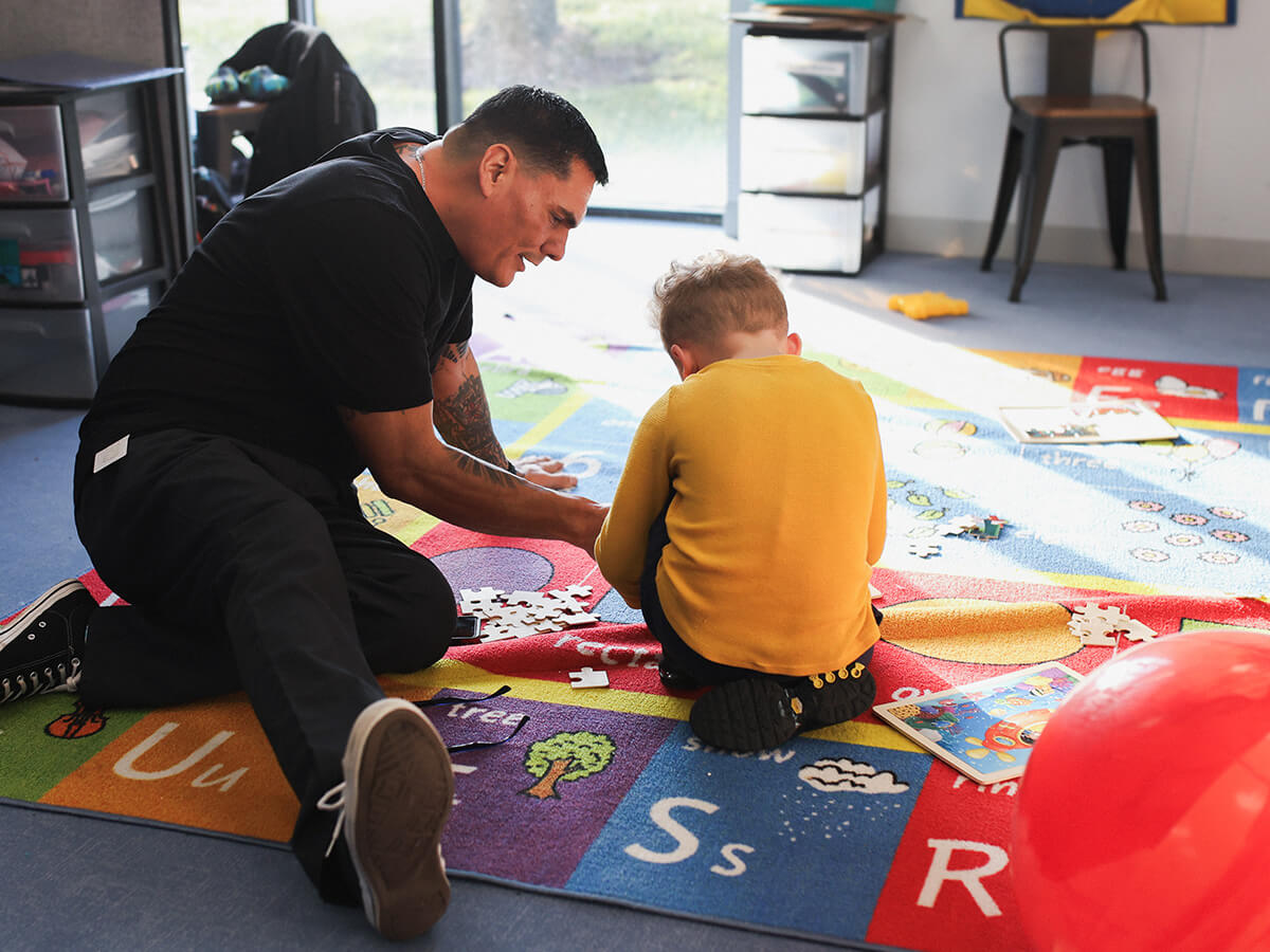 Adult and child assembling a puzzle on a colorful alphabet mat in a bright playroom.