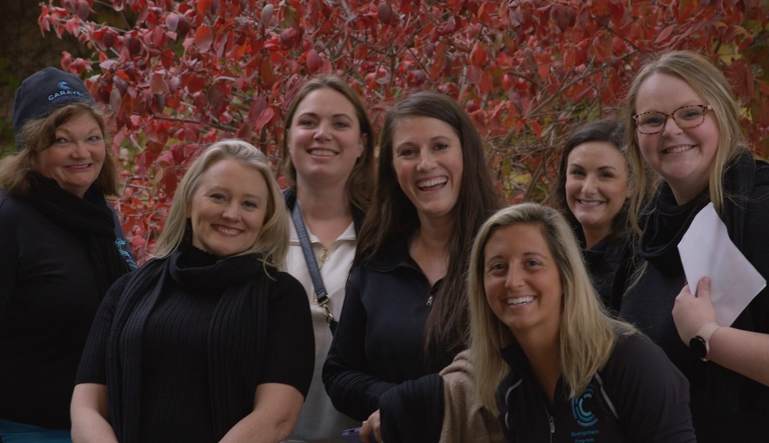 Group of seven women smiling with red autumn leaves in the background.