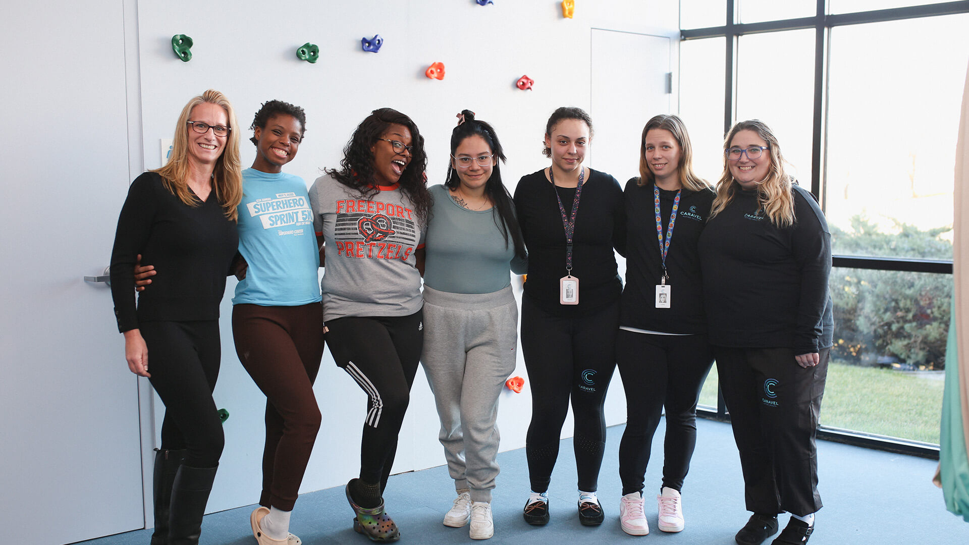 Seven women smiling and posing together indoors by a climbing wall.