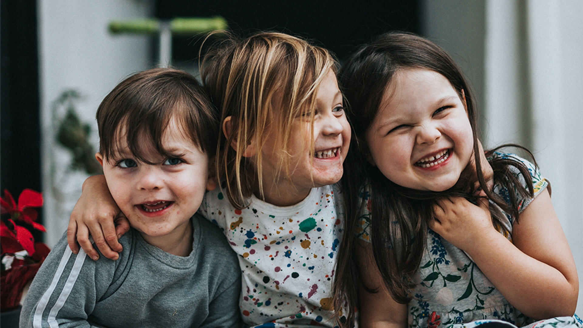 Three smiling children sitting close together, one with arm around another, indoors.