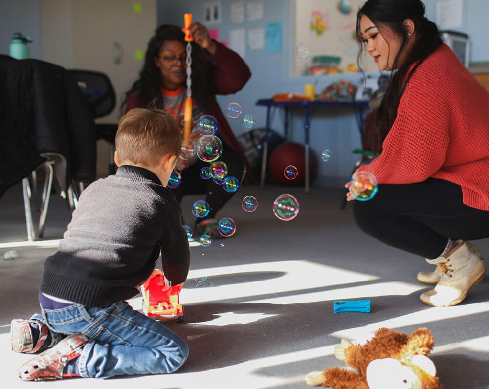 Child blowing bubbles with teachers during an interactive play session.