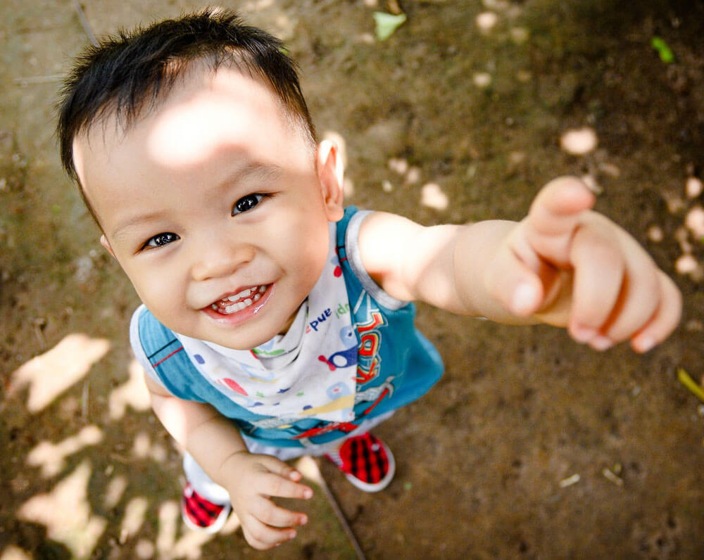 Joyful baby boy reaching up with a big smile, standing on a sunny outdoor path.