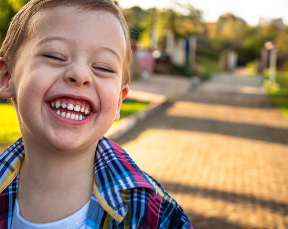 Young boy smiling with eyes closed on a sunny pathway, wearing a plaid shirt.