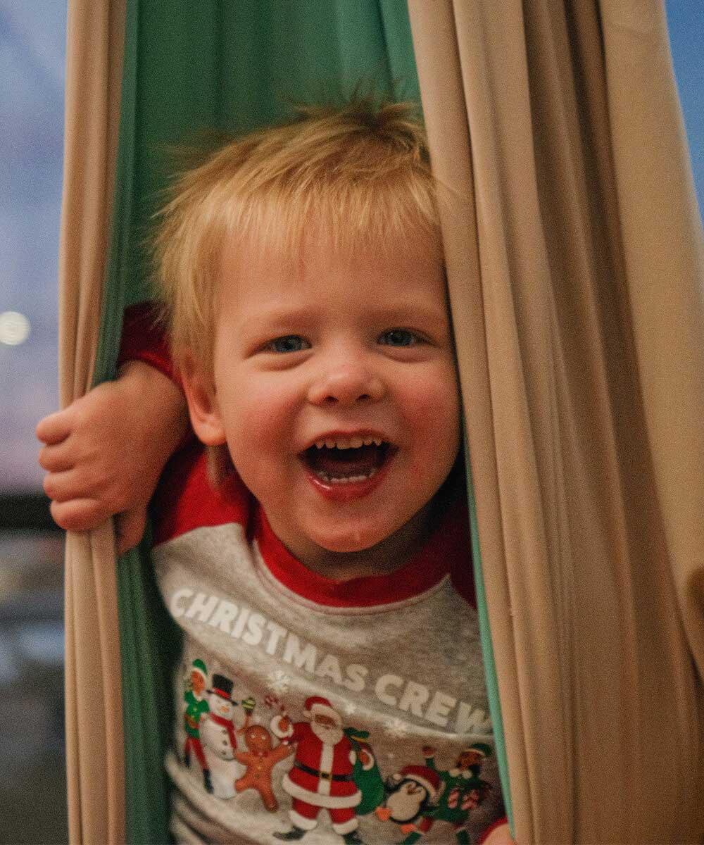 Smiling child in Christmas pajamas peeking out from curtains with joy.