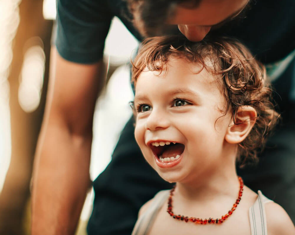 Young child with curly hair smiling brightly outdoors wearing a beaded necklace.