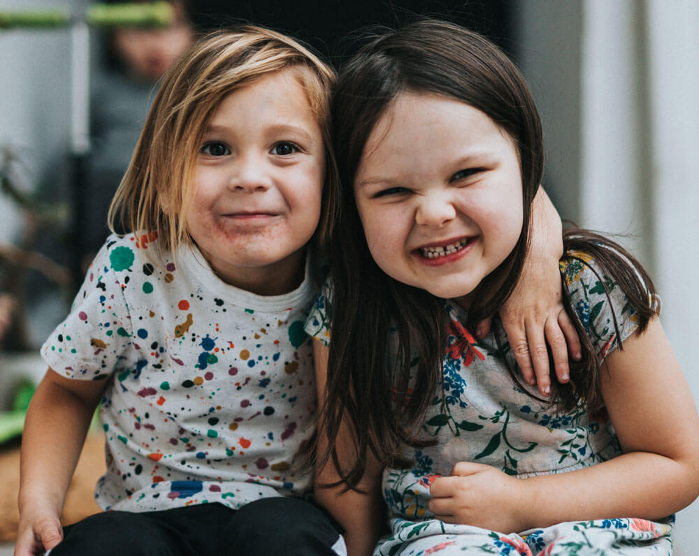 Two young girls sitting together, smiling with arms around each other indoors.