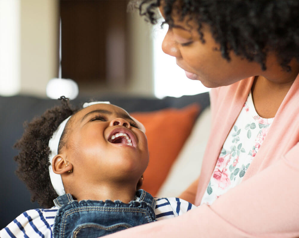 Smiling young girl in overalls embraced by a caring parent indoors.