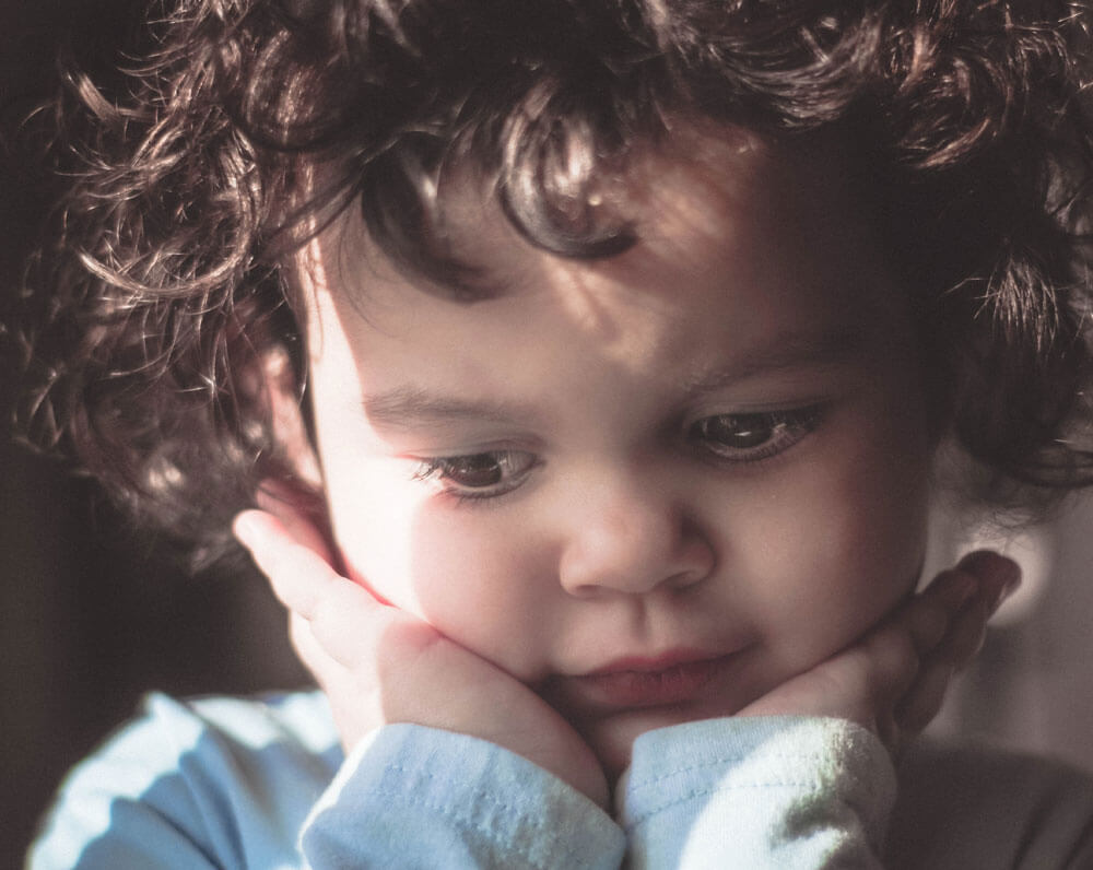 Young child with curly hair resting head on hands in soft lighting.