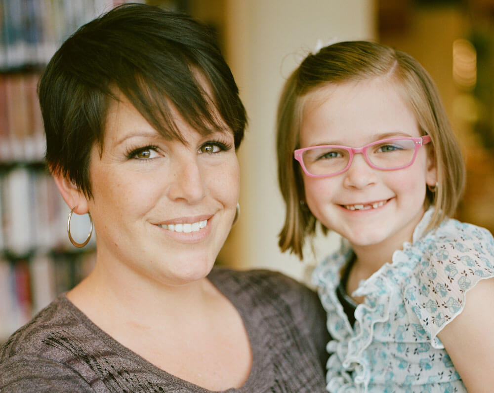 Smiling woman with short hair and young girl with pink glasses indoors.