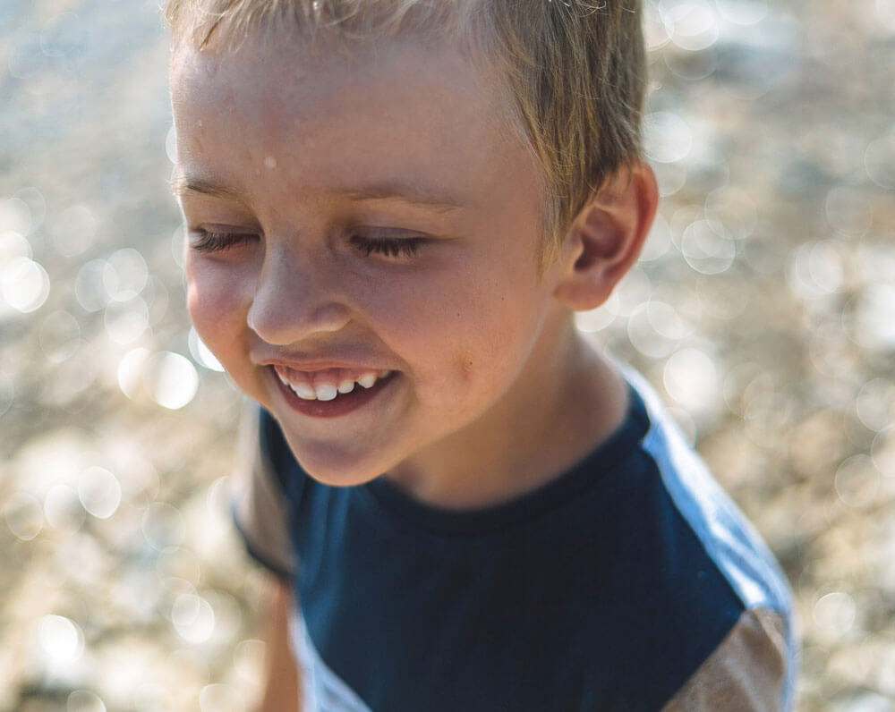 Young boy with closed eyes smiling warmly in bright sunlight outdoors.