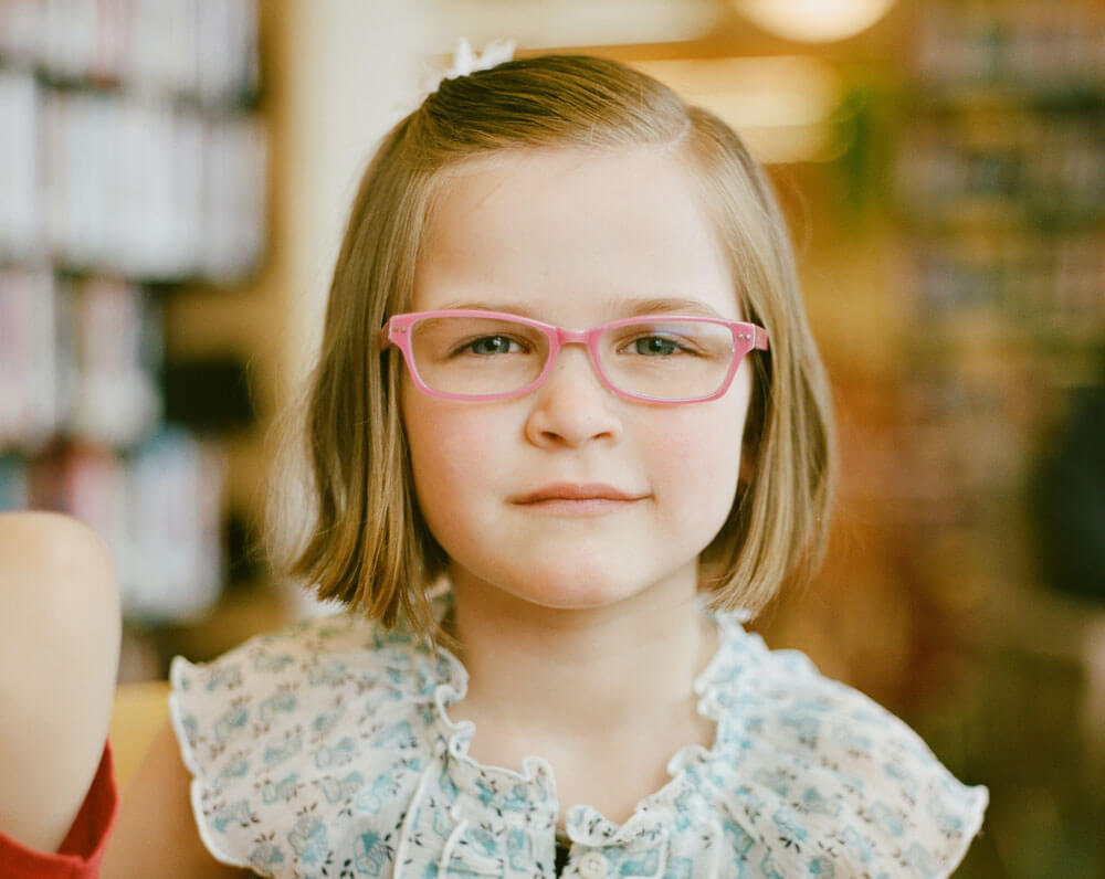 Young girl with short hair and pink glasses in a light blue blouse indoors.